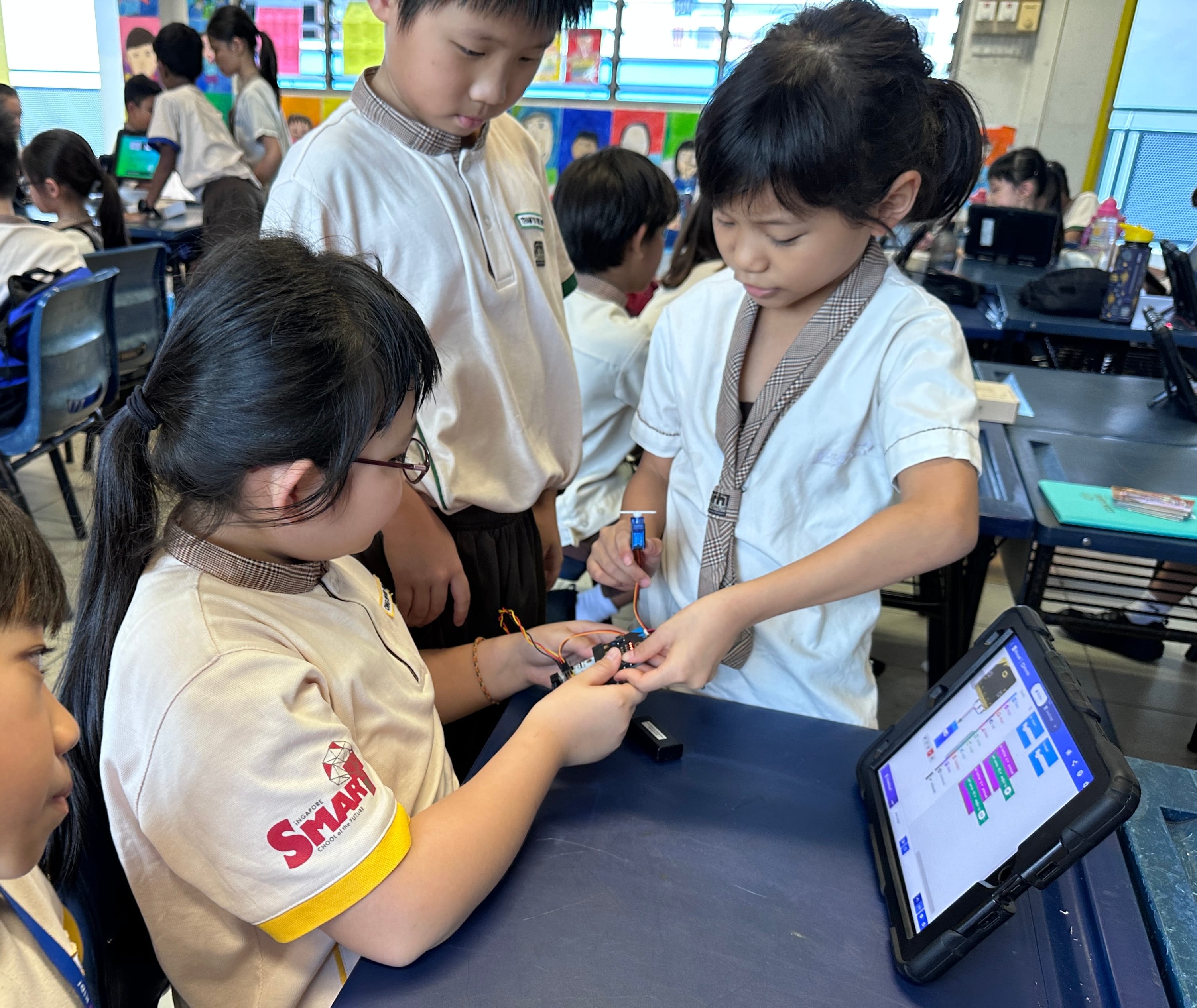 Three young students in school uniforms work together on a small electronic circuit next to a tablet showing a coding interface.